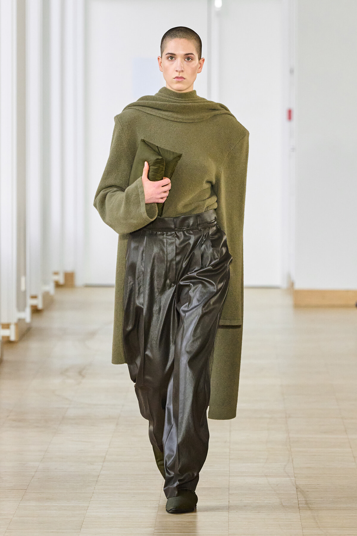 Model with a shaved head walks a runway wearing an olive green oversized sweater and black shiny leather pants, clutch in hand in a neutral hallway setting.