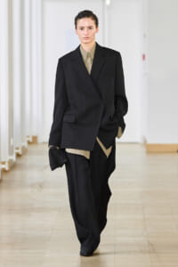 Model walks a runway in an oversized black suit with a beige shirt, hands in pockets, minimalist backstage setting