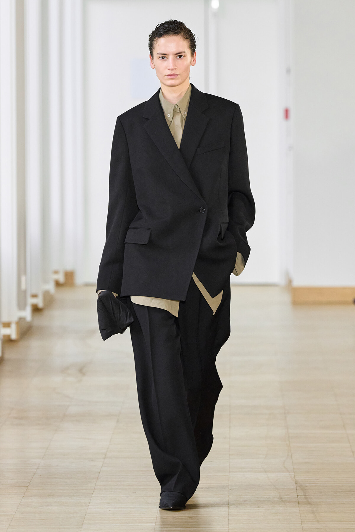 Model walks a runway in an oversized black suit with a beige shirt, hands in pockets, minimalist backstage setting