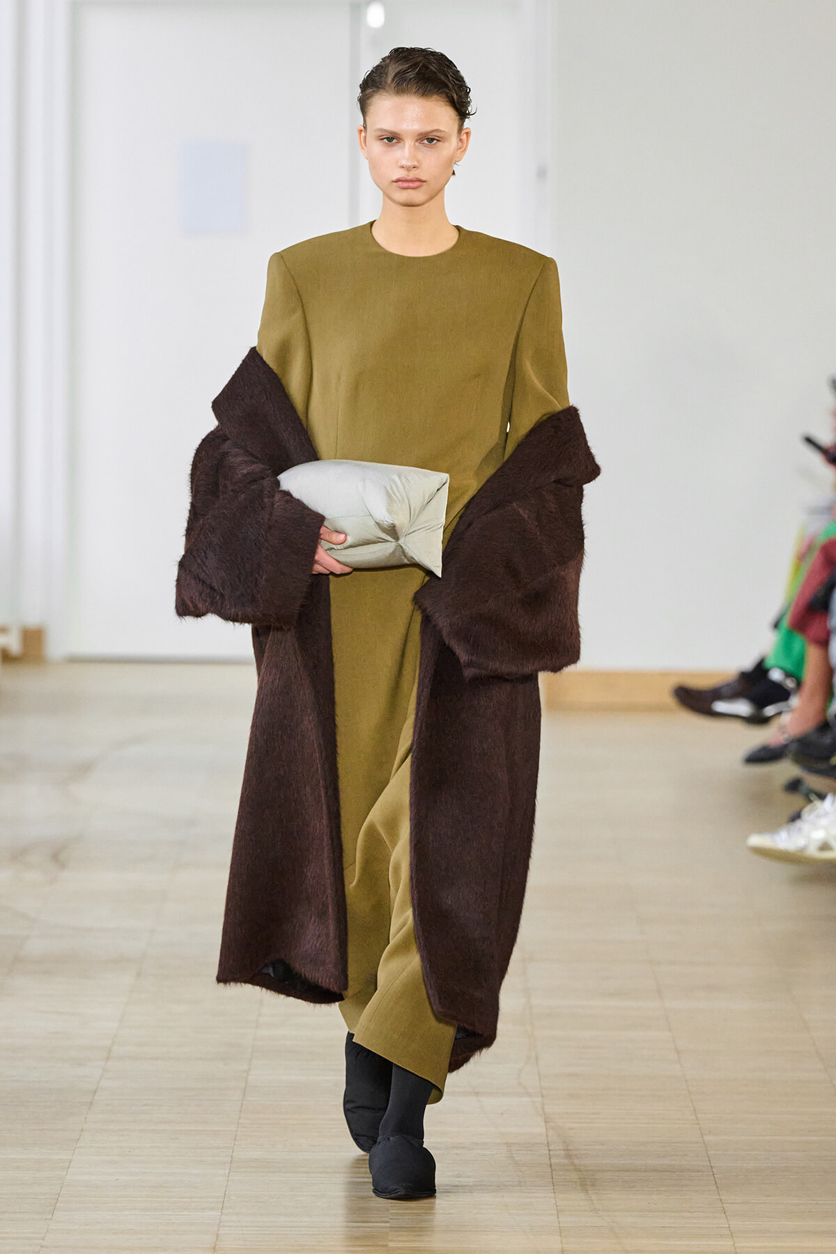 Model walking a runway in an olive-green dress with a dark brown oversized fur coat, holding a light gray rectangular clutch.