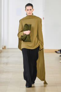 Runway model in an oversized olive cape top with black wide-leg pants, holding a dark green clutch as she walks the catwalk.