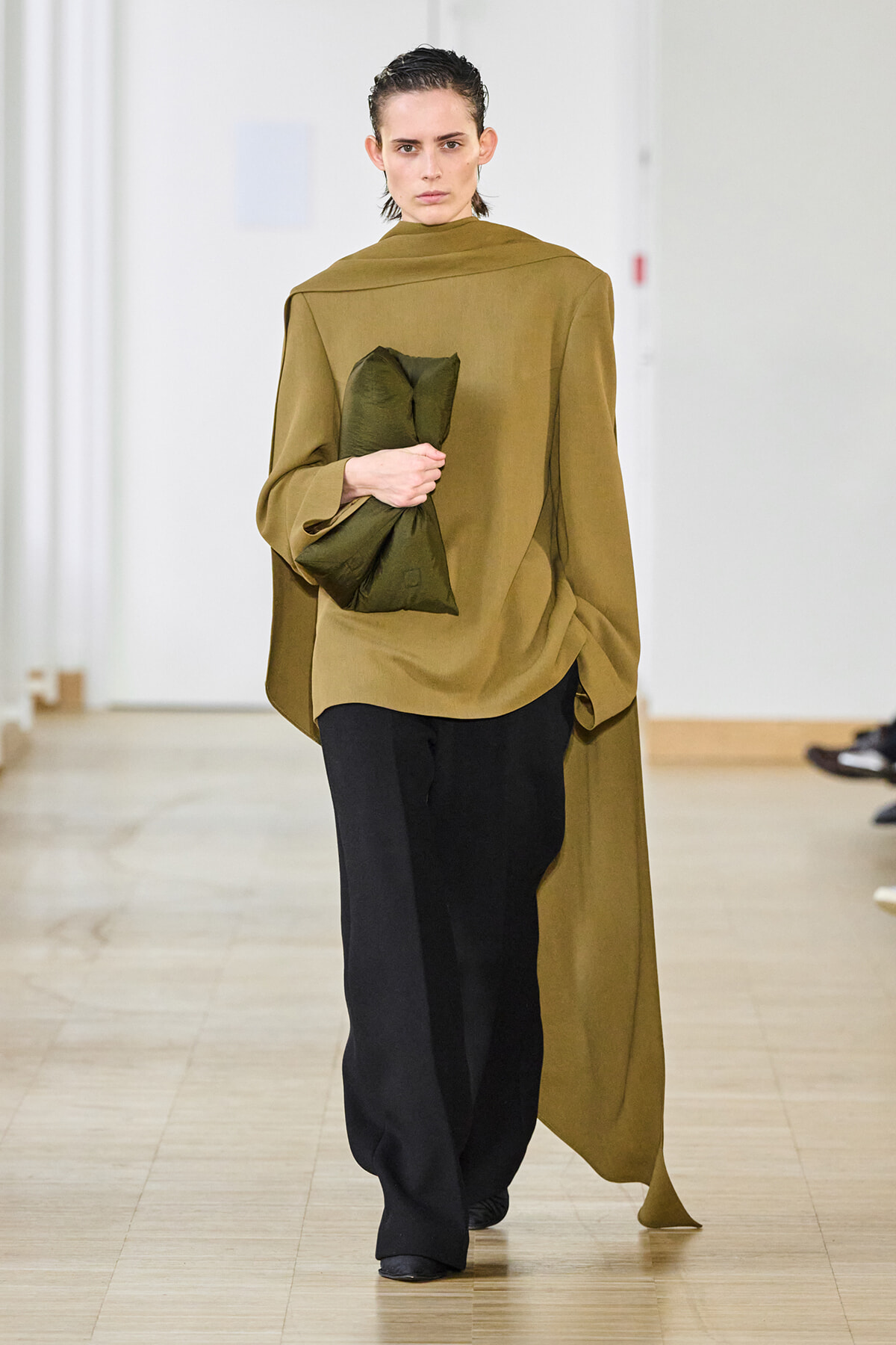 Runway model in an oversized olive cape top with black wide-leg pants, holding a dark green clutch as she walks the catwalk.