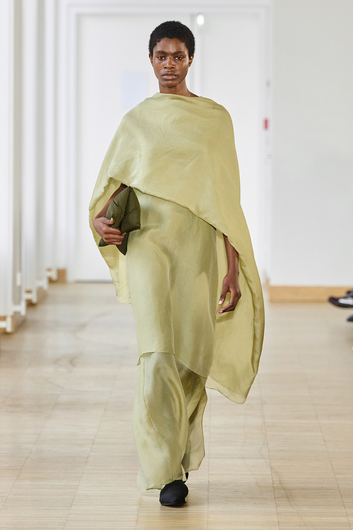 Model walking a runway in a pale olive draped cape-style dress, holding a dark green clutch in front of her, with black shoes and a neutral backdrop.