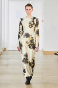 Model walks down the runway in a cream satin ankle-length dress with long sleeves and black abstract floral prints on a pale background.