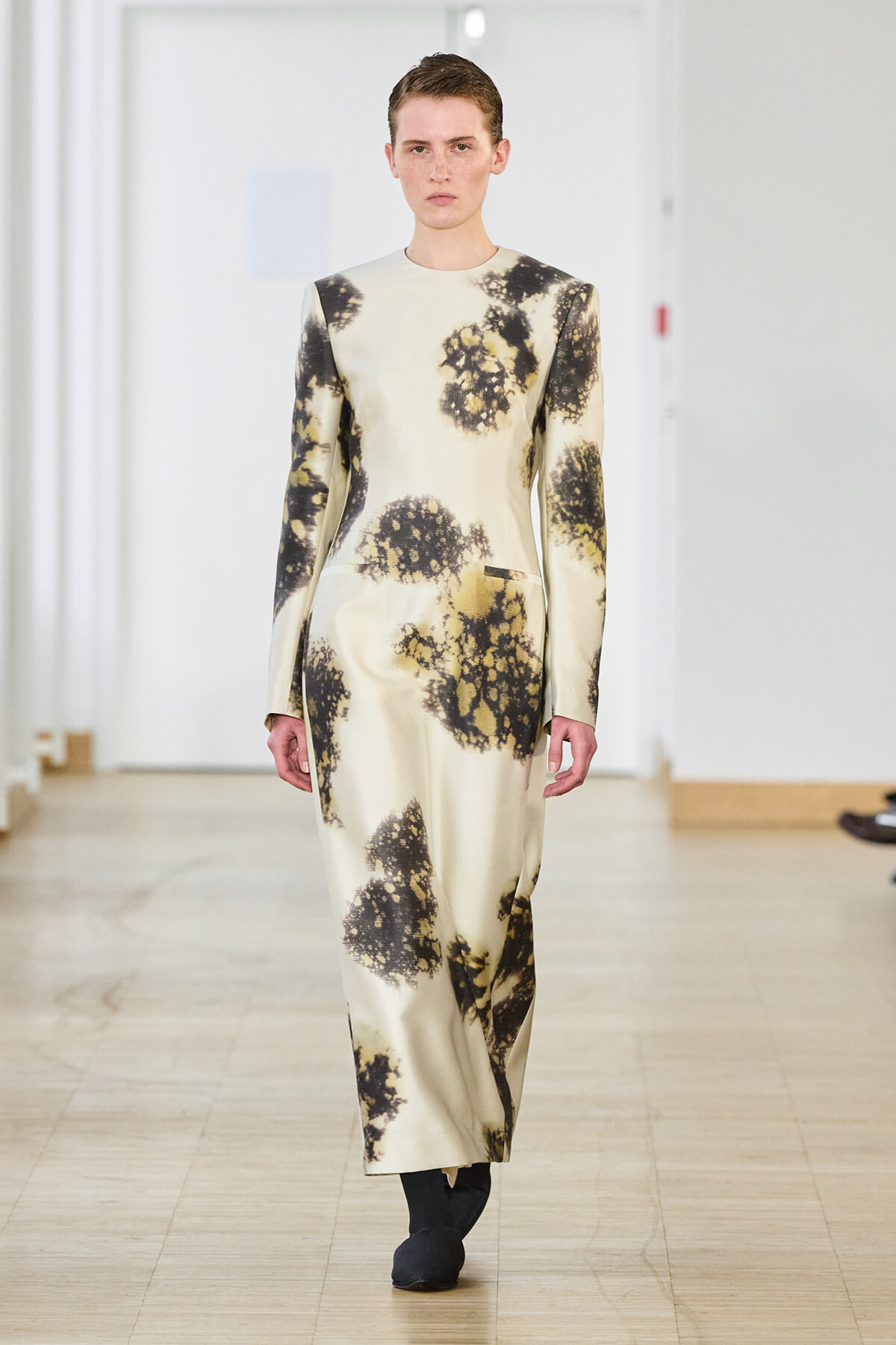 Model walks down the runway in a cream satin ankle-length dress with long sleeves and black abstract floral prints on a pale background.