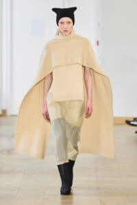 Model walks the runway in a large beige cape over a cream sweater and pale pants, paired with a black knit beanie and black ankle boots
