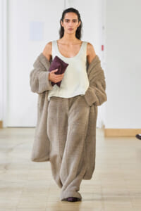 Model walking in an oversized beige teddy coat and matching wide pants, holding a burgundy clutch.