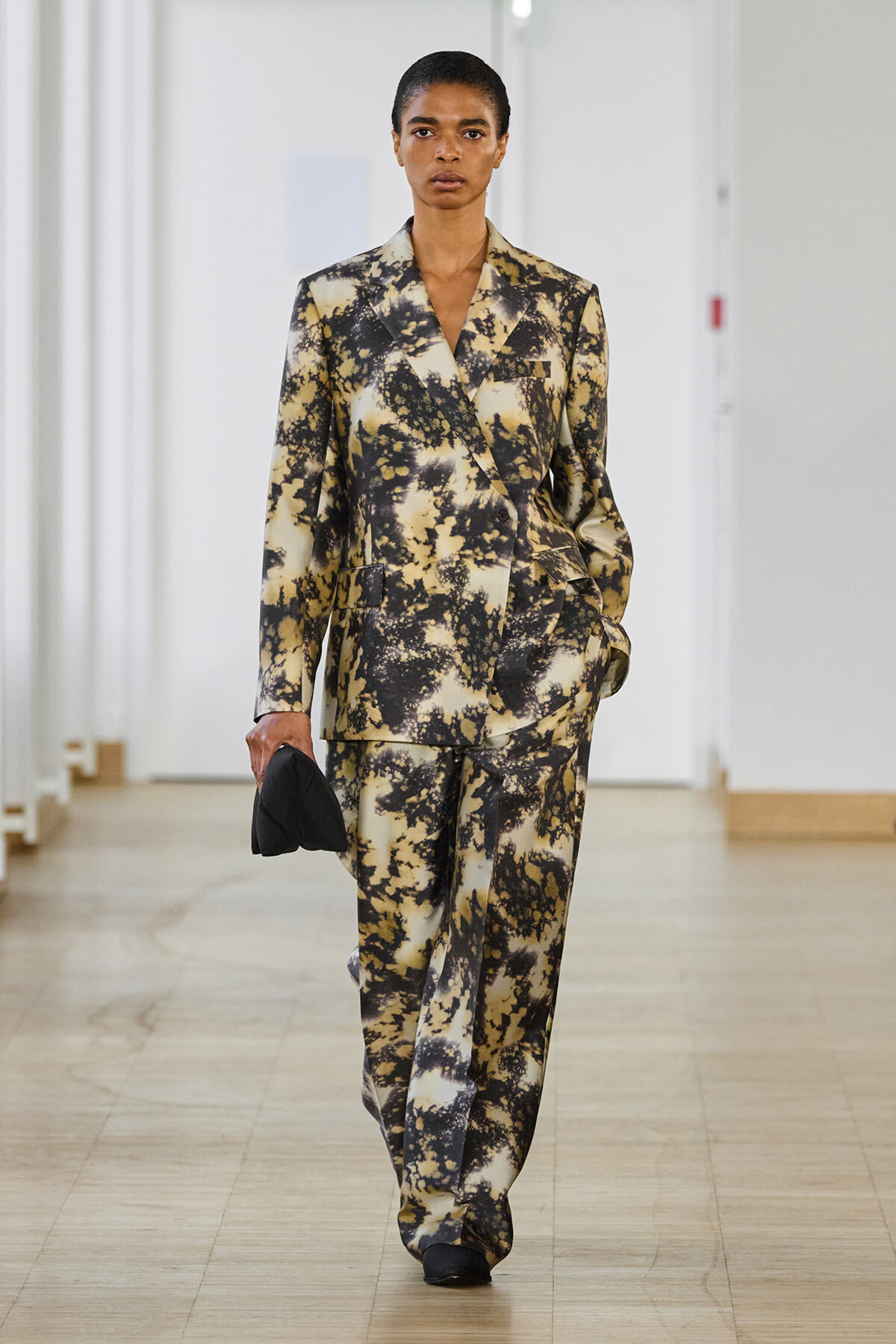 Model walking a runway in a beige and black abstract-print suit (oversized blazer and wide-leg pants), holding a small black clutch.