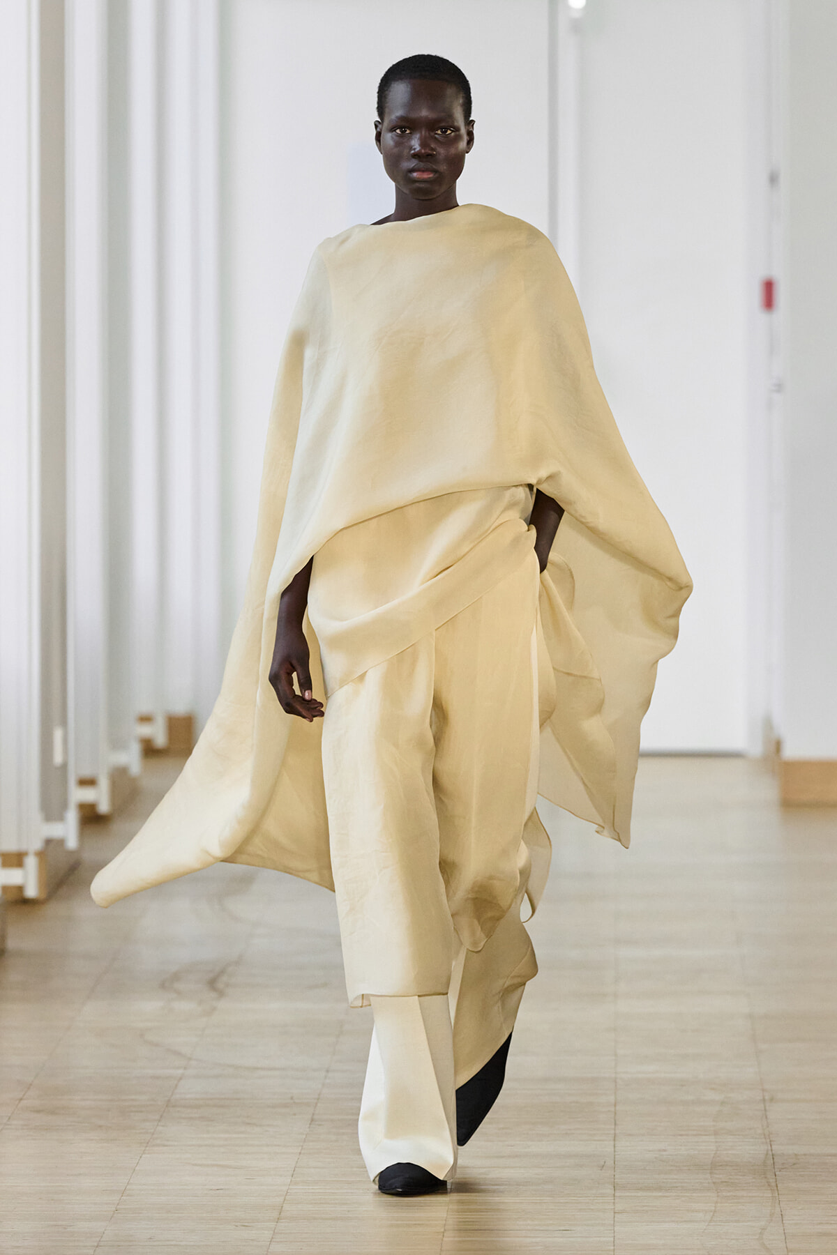Model walking in an oversized ivory cape and matching wide-leg pants on a runway.