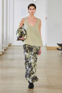 Model walking a runway in a neutral sleeveless top and tie-dye snake-print pants, holding a patterned scarf under her arm.