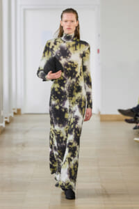 Model walking down a runway in a full-length tie-dye jumpsuit (cream, black, olive) while holding a black clutch and gazing forward.