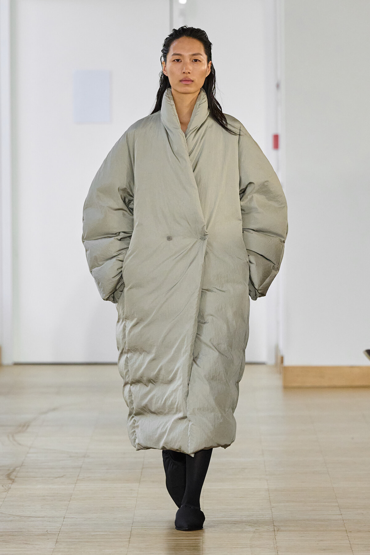 Model walking down a runway in an oversized pale gray puffer coat, full-body view, neutral expression.