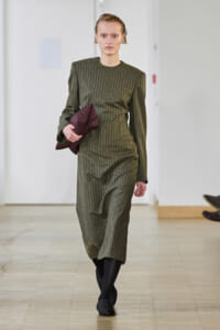 Model walking a runway in an olive-green pinstriped midi dress, carrying a burgundy jacket under her arm.