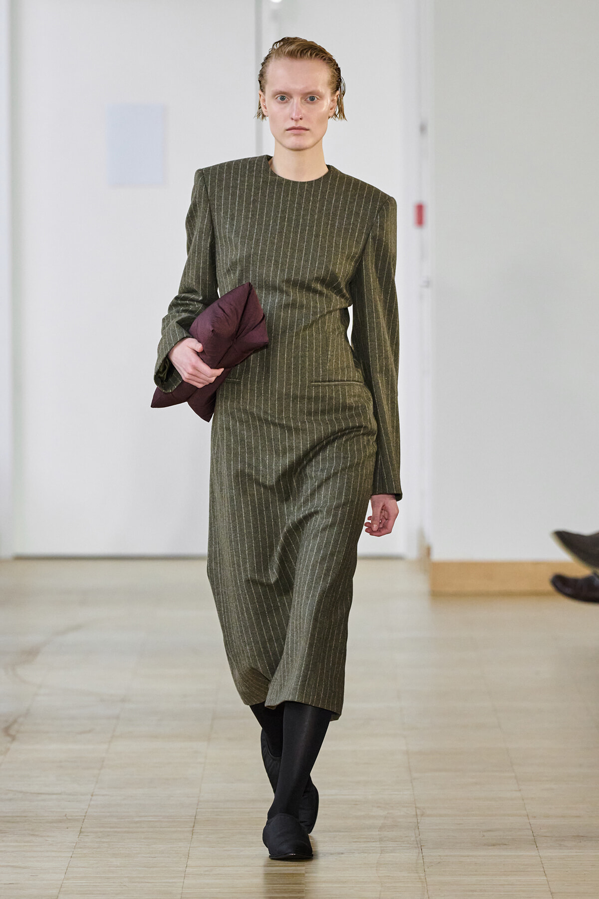 Model walking a runway in an olive-green pinstriped midi dress, carrying a burgundy jacket under her arm.