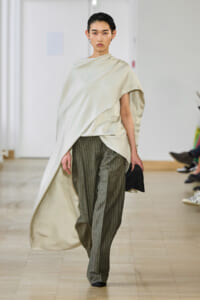 Model walking a fashion runway in an oversized cream draped top and gray pinstriped wide-leg pants, holding a black clutch at her side.