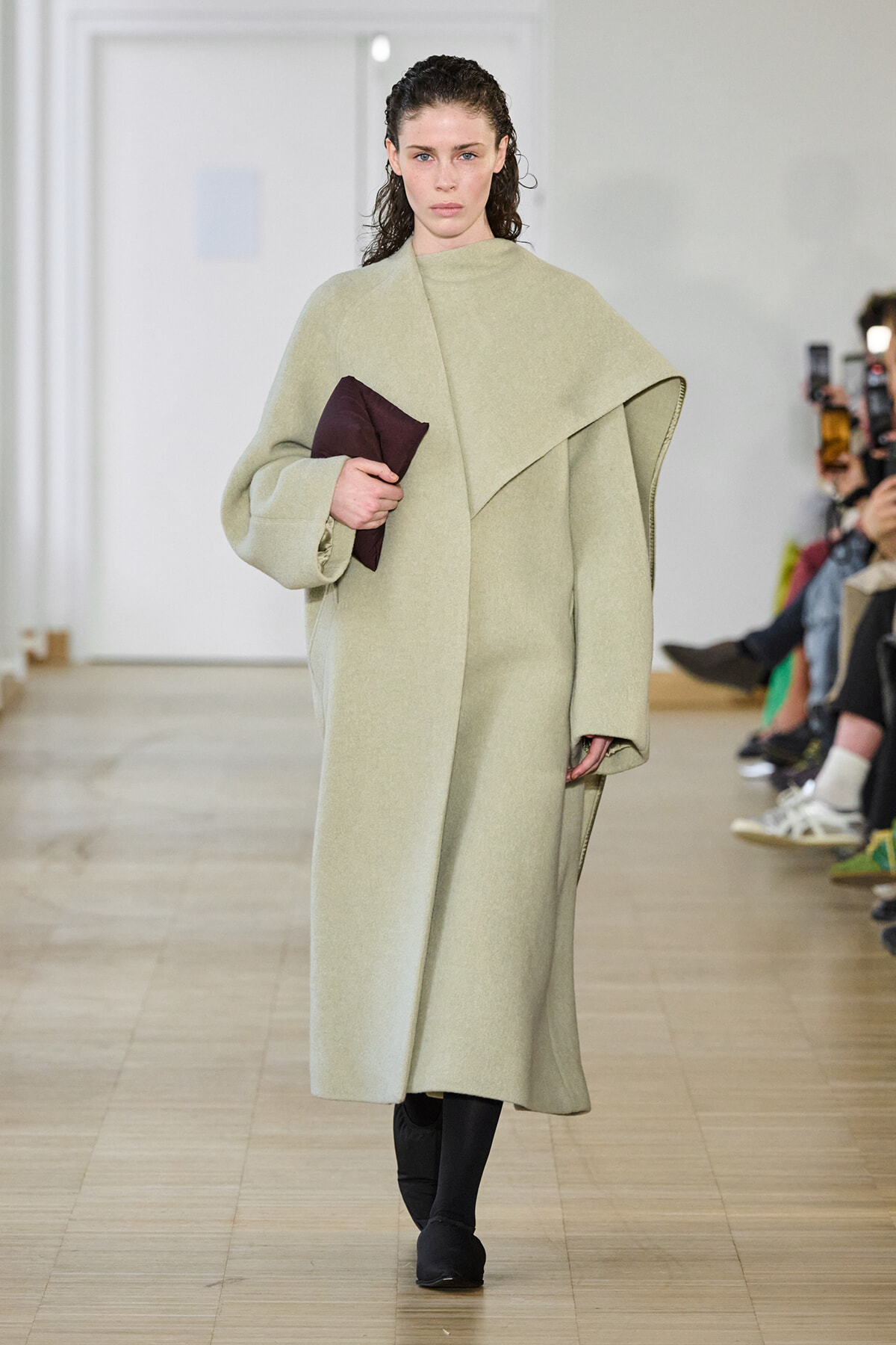 Model walking a runway in a pale beige oversized coat, holding a dark burgundy clutch; audience in the background