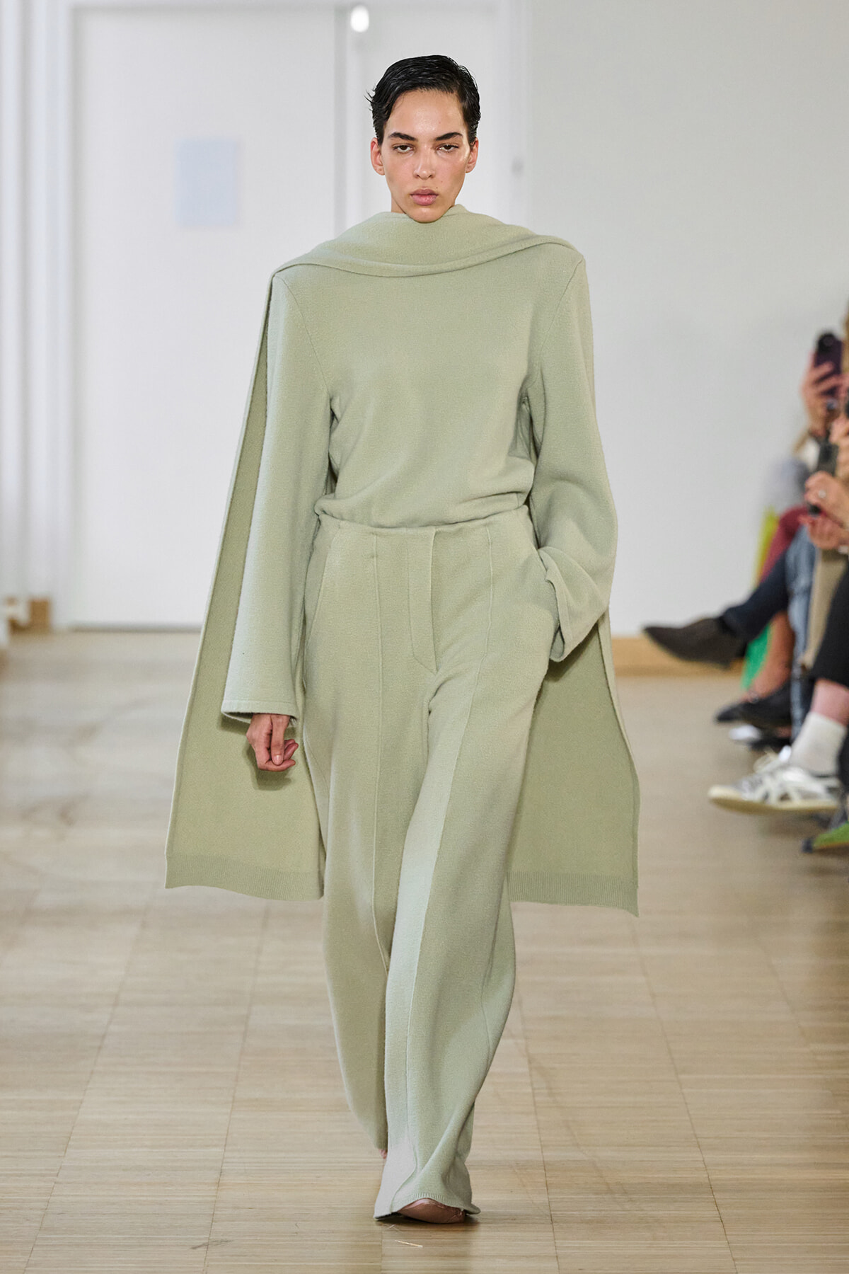 Model walking the runway in a pale sage matching cape top and wide-leg trousers on a fashion show runway.