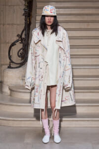 Model in a cream, signature-covered coat with a matching bucket hat, pink leg warmers, and white shoes, standing on stone stairs.