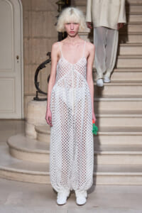 Model walking a runway in a sheer white grid-pattern dress with thin straps, standing on a staircase during a fashion show, holding a small bright green accessory.