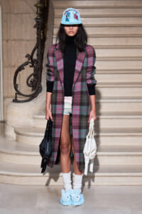 Model on stone stairs wearing a long pink-gray plaid coat over a black turtleneck and white shorts, with white socks, light-blue cap, fluffy blue bunny slippers, and two handbags.