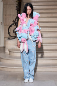 Model wearing a pink and blue plush bunny fur wrap over a pale outfit, with light blue star-patterned jeans and white shoes on a stairway set.