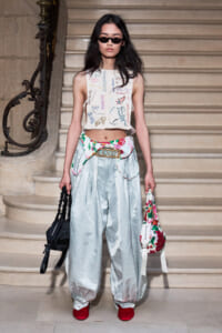 Model on a stone staircase wearing a white cropped top with colorful signatures, silver baggy pants, red shoes, sunglasses, and a floral bag.