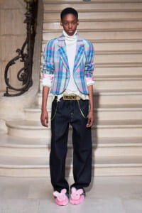 Model on a staircase runway wearing a pastel plaid blazer over a white turtleneck, loose dark jeans with neon-green accents, a gold Kenzo belt, and pink fluffy bunny slippers.