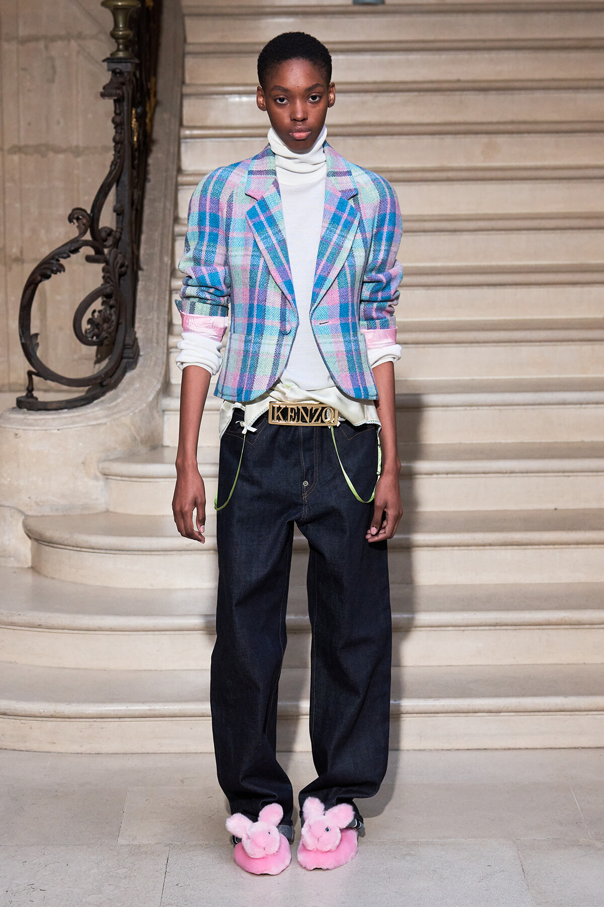 Model on a staircase runway wearing a pastel plaid blazer over a white turtleneck, loose dark jeans with neon-green accents, a gold Kenzo belt, and pink fluffy bunny slippers.