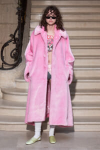 Model in an oversized pink fur coat on a stairway, wearing black sunglasses, a graphic top, white socks, and pale green flats.