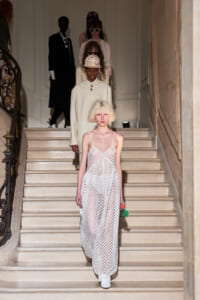 Model in a white mesh maxi dress descends a grand staircase during a fashion show; background models follow on the stairs.