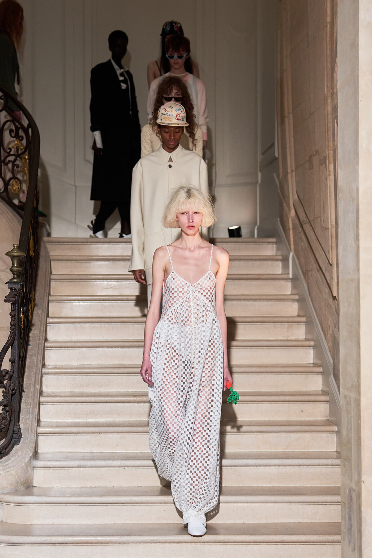 Model in a white mesh maxi dress descends a grand staircase during a fashion show; background models follow on the stairs.