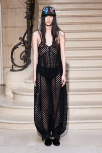 Model in a black checkered mesh dress standing on a staircase, wearing a cap with colorful patches.