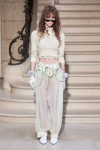 Model on a grand staircase wearing a cream crochet crop top, sheer ivory pants, white gloves, fur cuffs, and sunglasses.