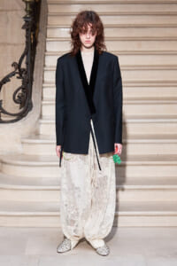 Person on a grand staircase wearing a black blazer with velvet lapels, sheer top, and wide lace pants, holding a green accessory.