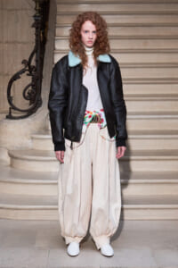 Person with curly red hair wearing a black leather jacket with light blue collar, white turtleneck, and oversized cream pants on a staircase backdrop, white shoes visible