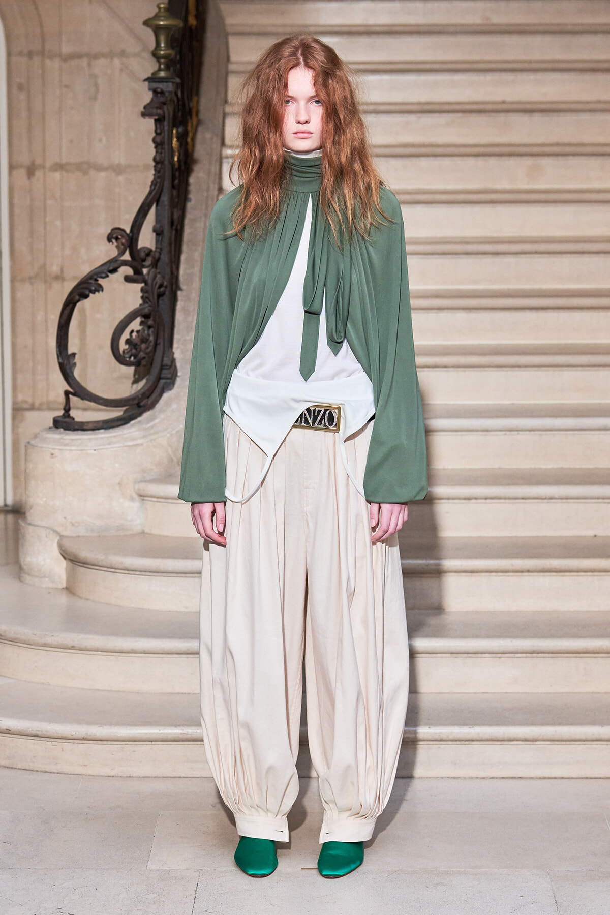 Model on a staircase wearing a green cape top, cream oversized pants, and green shoes for a fashion show.