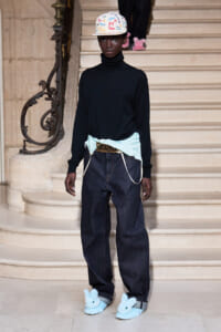 Model stands on a grand staircase in a black turtleneck, loose dark jeans, a light blue wrap belt with cords, a signed white cap, and blue bunny slippers.