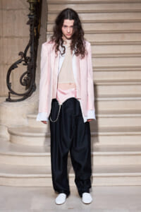 Model on a stone staircase wearing a pale pink blazer over a sheer beige top and oversized black pants with white shoes.