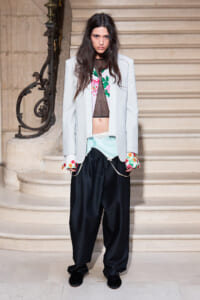Model standing on a grand staircase in a pale blazer with floral cuffs, sheer black top, and loose black pants.