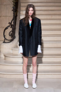 Model standing on a stone staircase in an oversized black blazer, with white shirt cuffs peeking out, pink knit socks, and white flats.