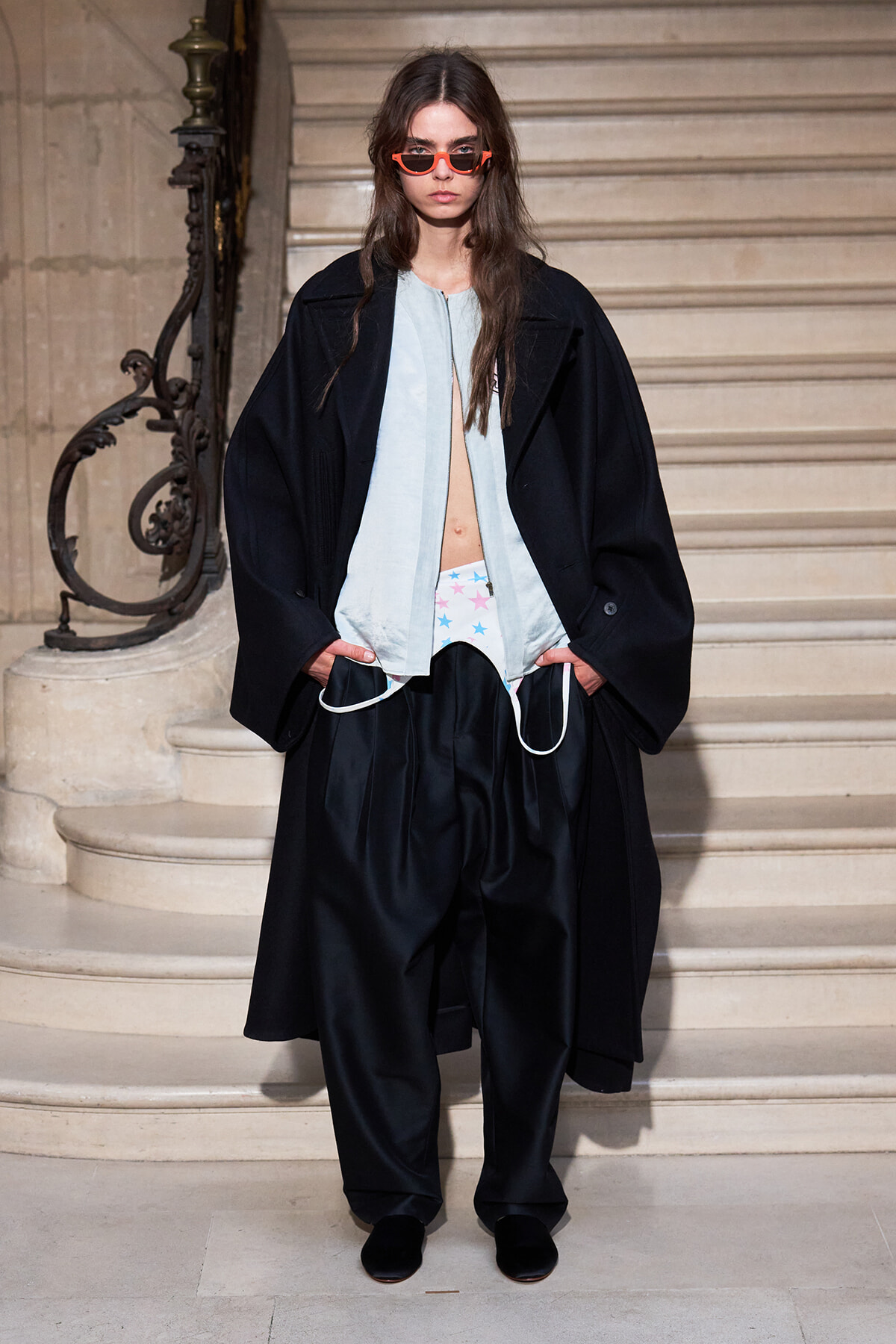 Model on a grand staircase wearing an oversized black coat, pale blue top, star-pattern underwear, wide black pants, and orange sunglasses.