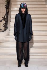 Person wearing a dark tailored blazer with velvet lapels, sheer black pants, and gloves, standing on a stone staircase wearing a embroidered cap with rainbow design.