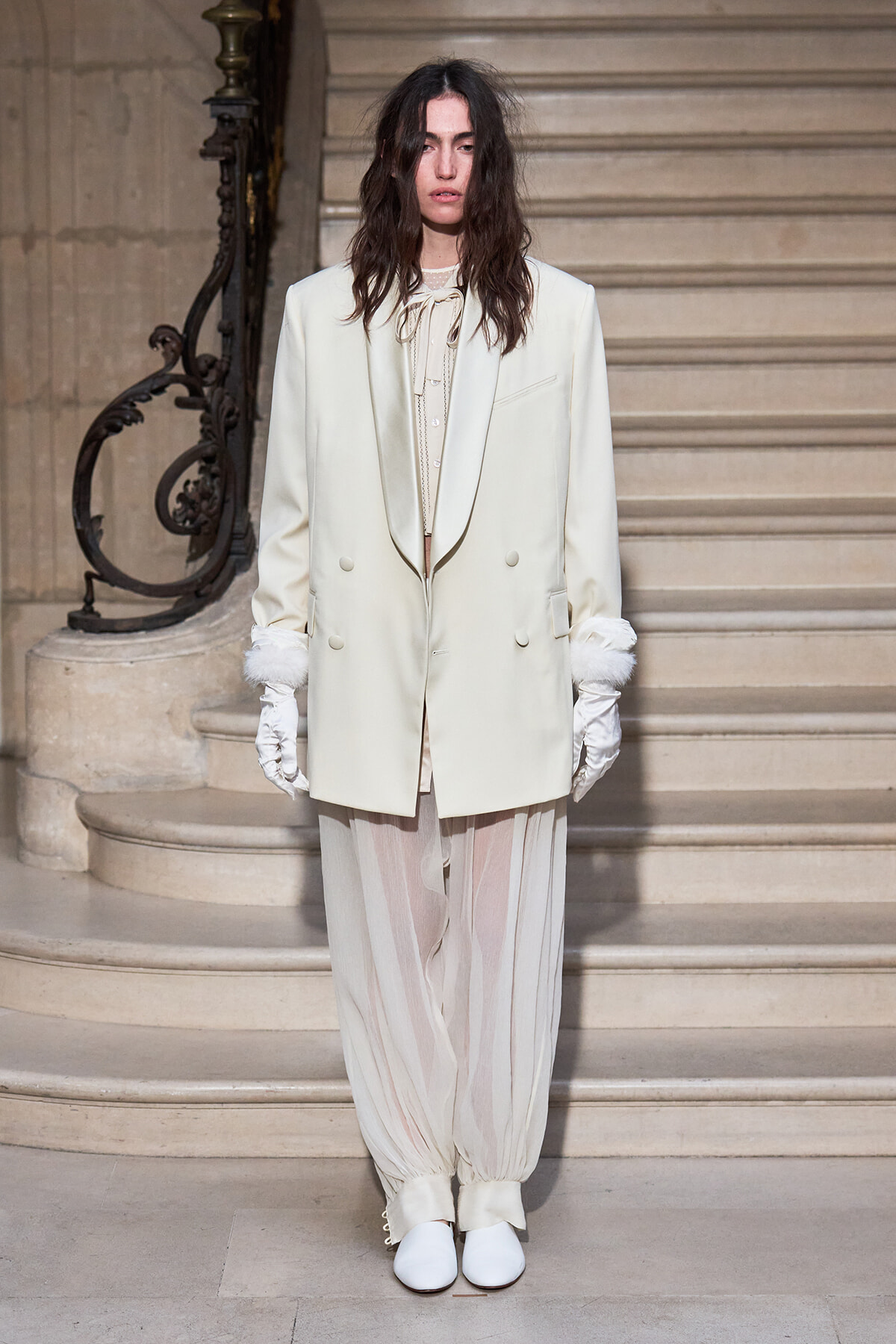 Model on a grand staircase wearing an ivory double‑breasted blazer, cream blouse, and sheer striped pants with white gloves and white shoes.