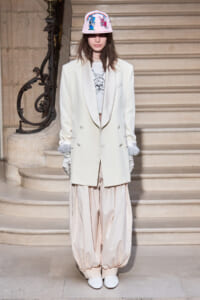 Model standing on a staircase in an oversized ivory blazer, pale balloon pants, white gloves, white shoes, and a pink cap featuring cartoon rabbits.