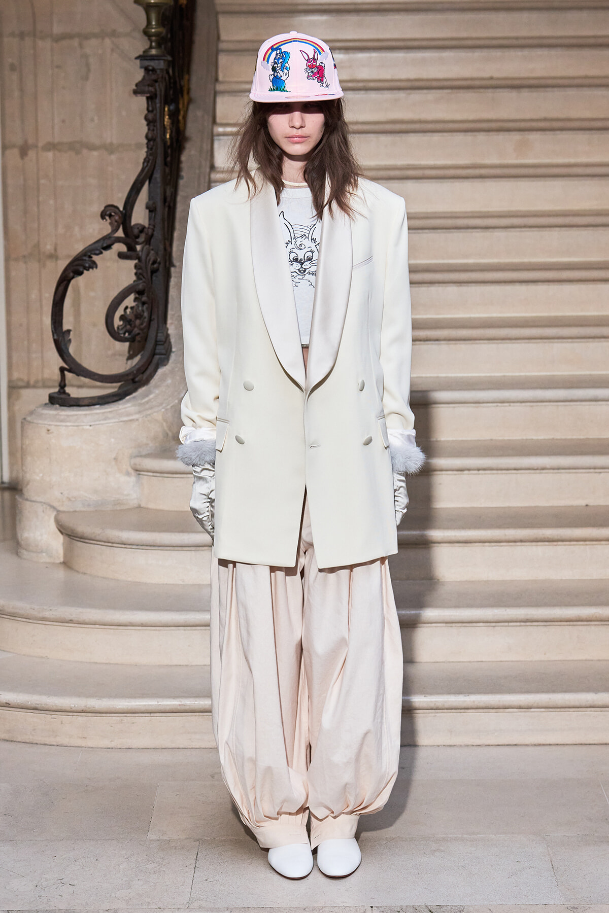 Model standing on a staircase in an oversized ivory blazer, pale balloon pants, white gloves, white shoes, and a pink cap featuring cartoon rabbits.