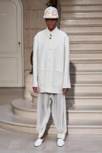 Model on a runway wearing an all-white ensemble: oversized coat with large front pockets, sheer gathered pants, white slip-on shoes, and a white cap covered in colorful signatures in front of a staircase backdrop.