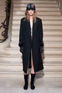 Model standing on a stone staircase in a long black double‑breasted coat with fur cuffs, black gloves, and a colorful embroidered cap; a patterned top shows at the chest.