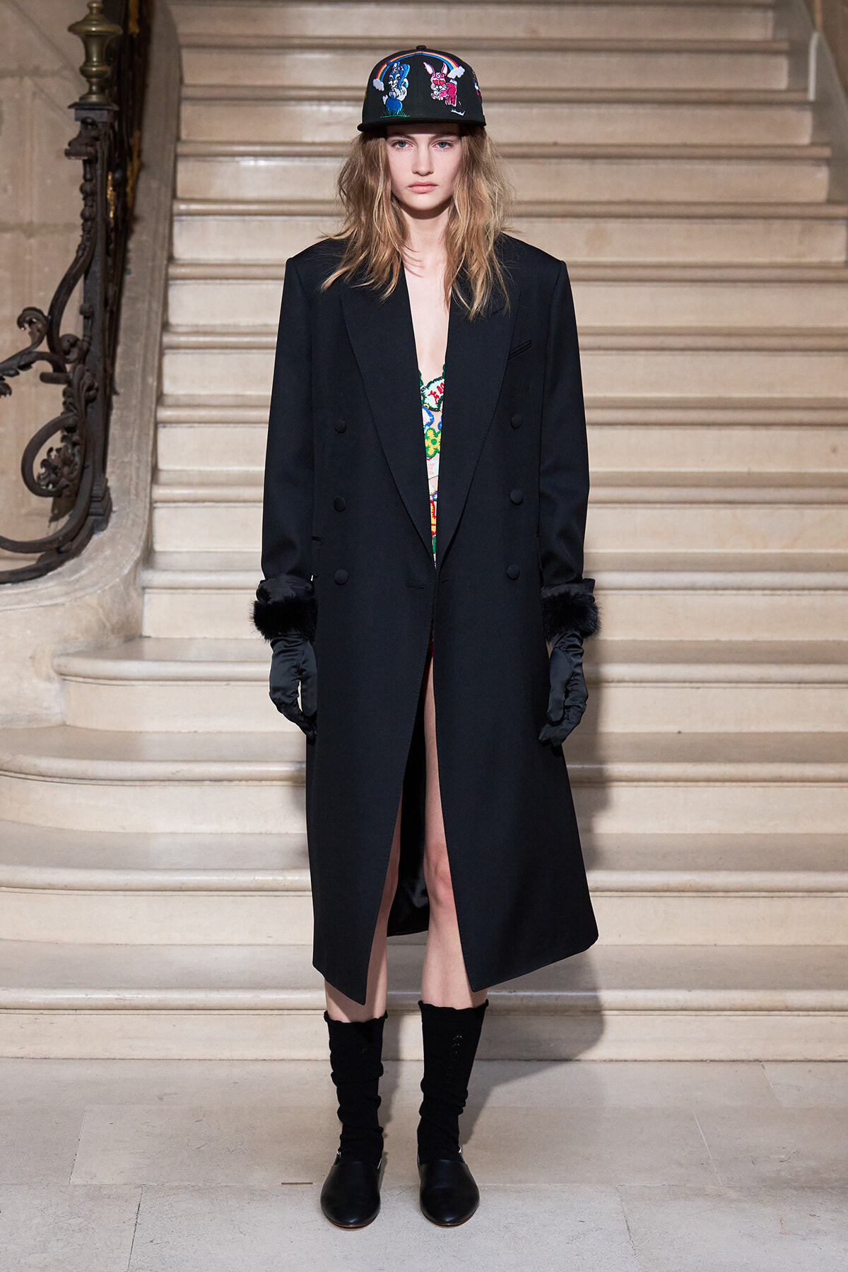 Model standing on a stone staircase in a long black double‑breasted coat with fur cuffs, black gloves, and a colorful embroidered cap; a patterned top shows at the chest.