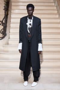 Model on a runway wearing a long black double-breasted coat with oversized white shirt cuffs, sheer black top, baggy black pants, and white slip-on shoes on a marble staircase.