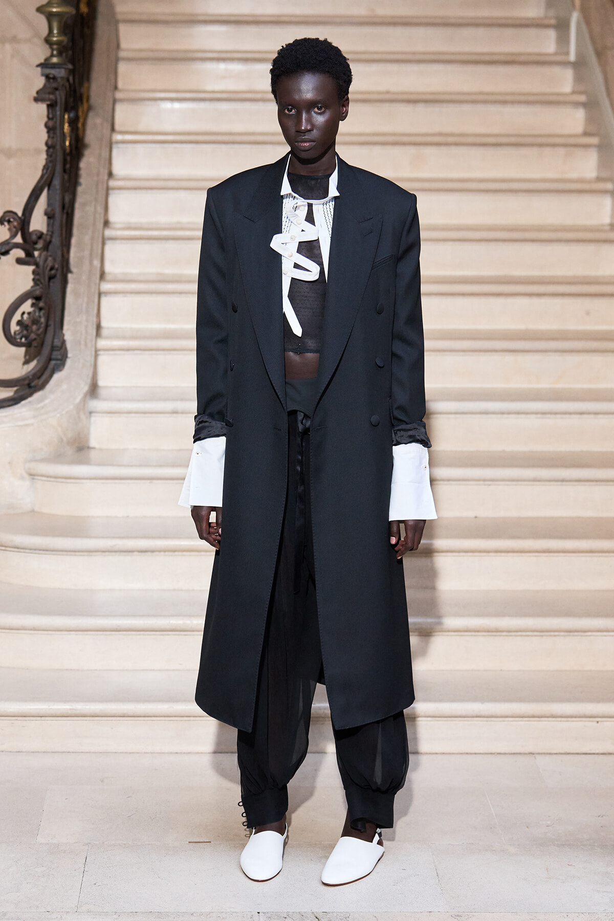 Model on a runway wearing a long black double-breasted coat with oversized white shirt cuffs, sheer black top, baggy black pants, and white slip-on shoes on a marble staircase.
